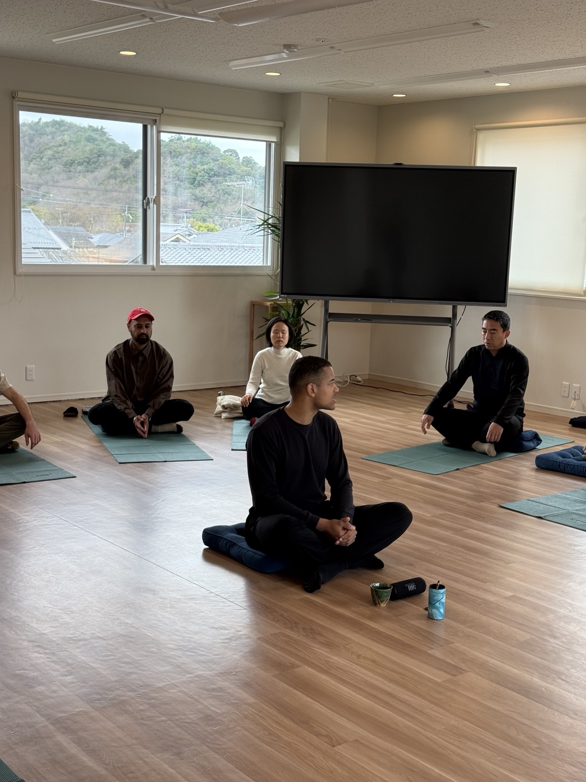 Retreat participants seated cross-legged on cushions and mats during a guided meditation, hills visible through the studio windows.