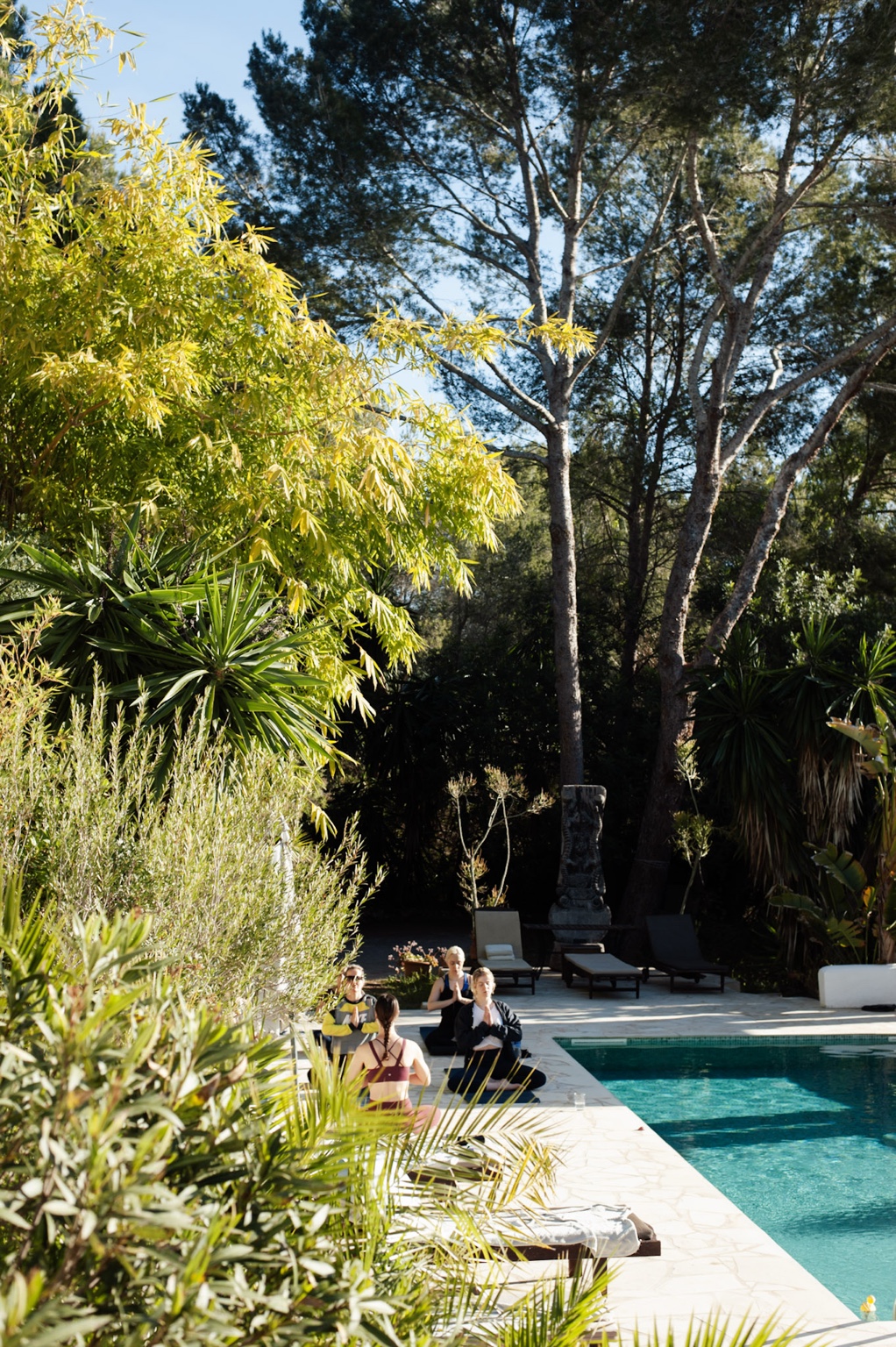 Small group seated in meditation on mats beside the villa pool in Ibiza, framed by tall pines and Mediterranean planting.