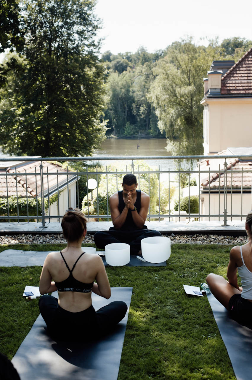 Nathan seated with crystal singing bowls leading a sound bath for participants on mats, with the lake and trees beyond the railing.