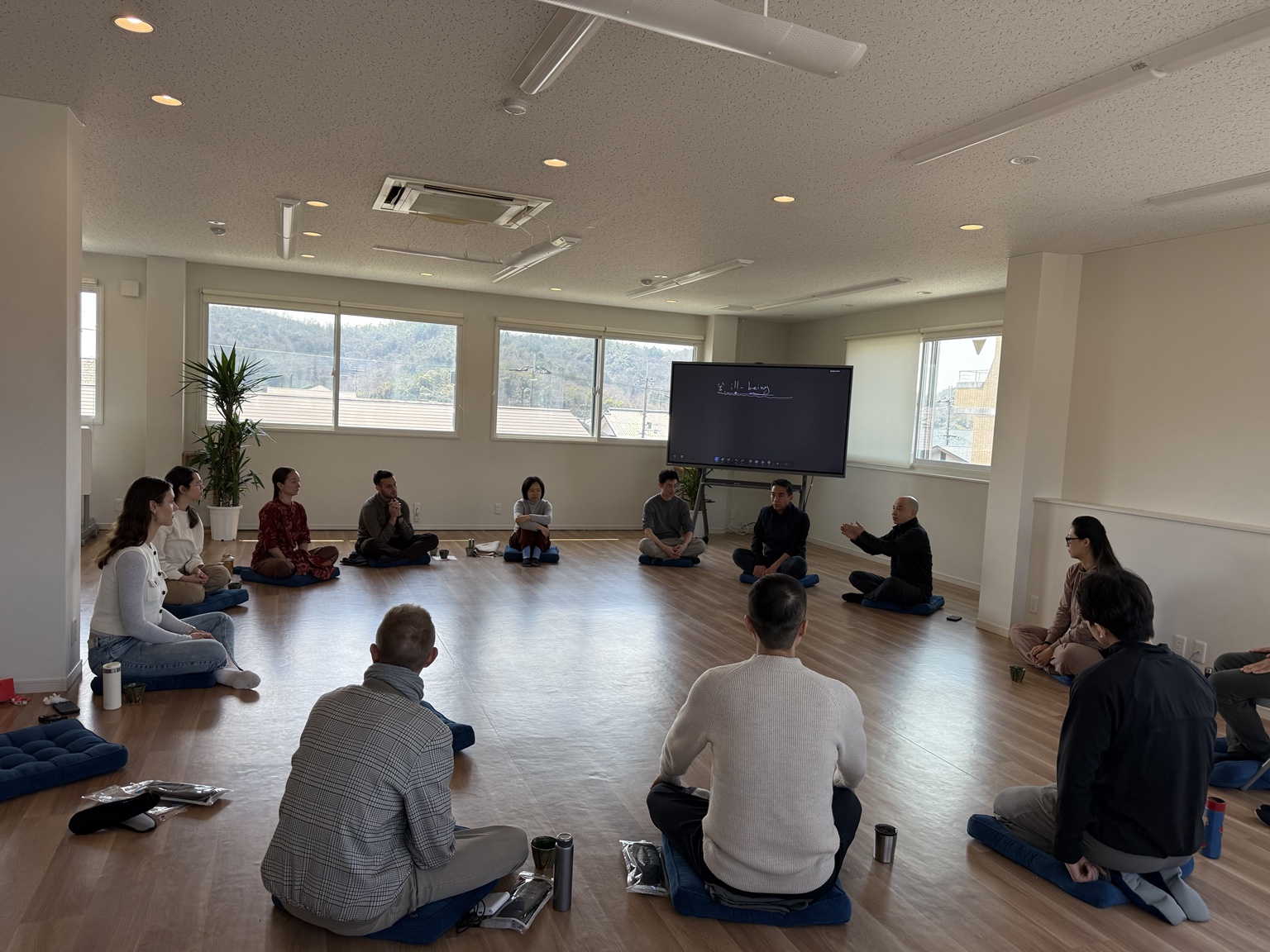 The full group seated in a wide circle on cushions for an open discussion, with wooded hills seen through the windows.