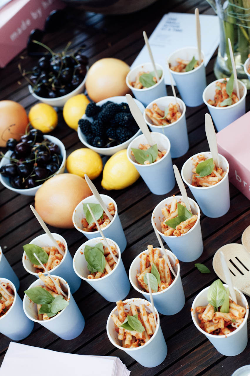 Cups of plant-based pasta topped with basil alongside bowls of cherries, blackberries, lemons and grapefruit on a wooden table.