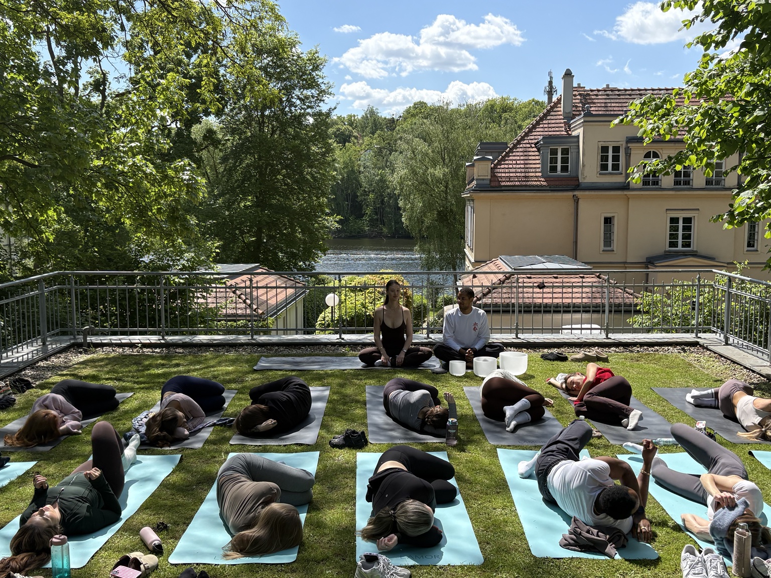 Outdoor group meditation on a lakeside terrace, participants resting on mats while Nathan and Kathi lead sound and reflection.