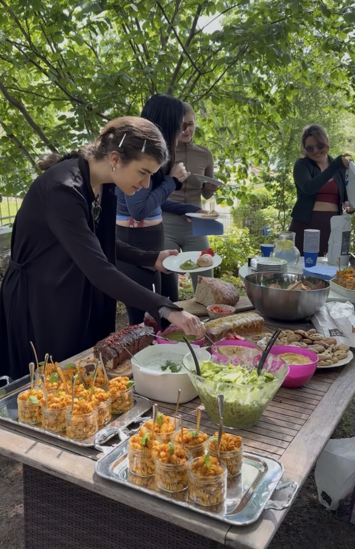 Guests serving themselves from a wooden table laid with plant-based salads, dips, breads and pasta jars under the trees.