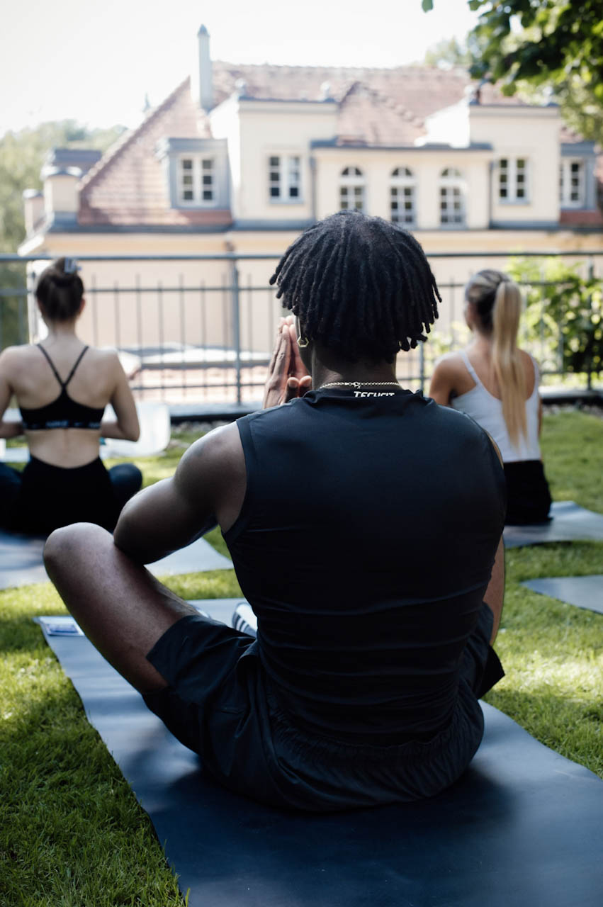 Participants seated with hands at heart centre during meditation on the garden lawn, facing a pale villa in the morning light.