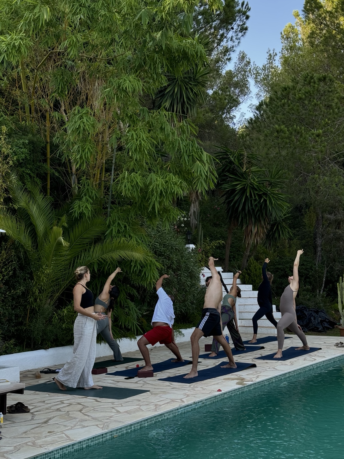 Participants in an extended side-angle stretch on mats beside the villa pool during a morning yoga class in Ibiza, framed by palms and pines.