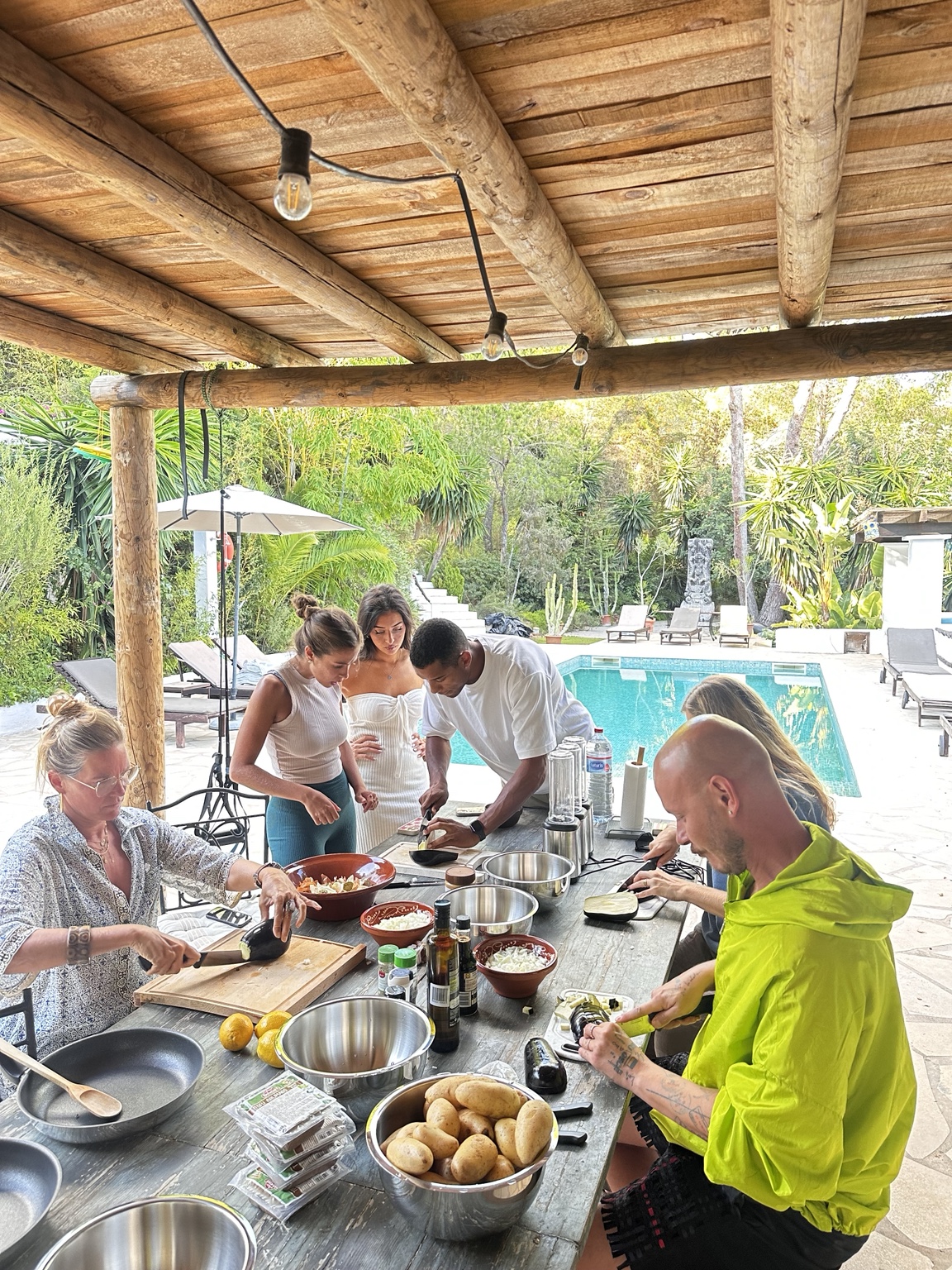 The group chopping vegetables together at a long wooden table under a timber pergola beside the pool during a mindful cooking session.