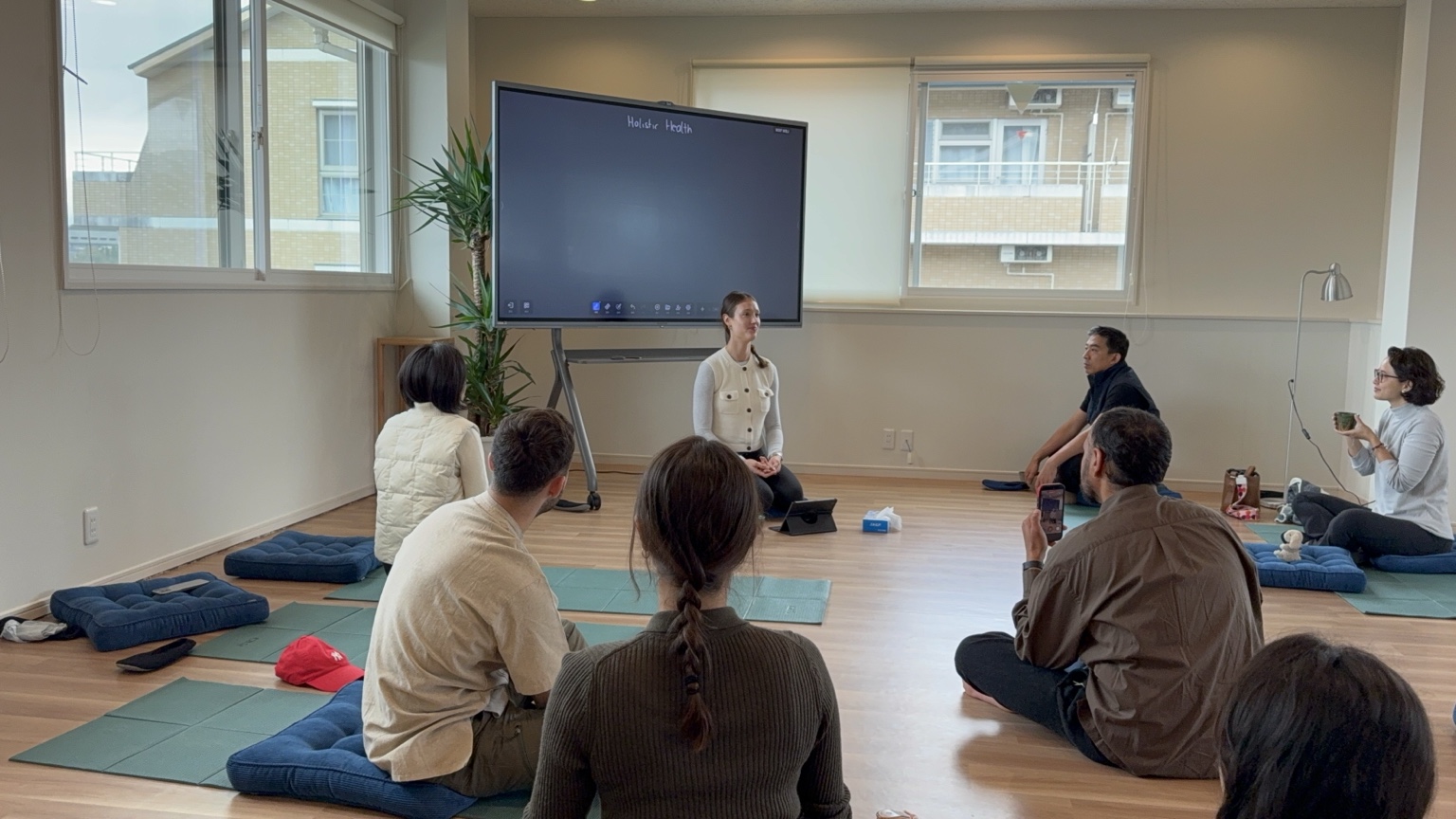 Katharina seated in front of a screen reading Holistic Health, leading a masterclass for the group seated on cushions.