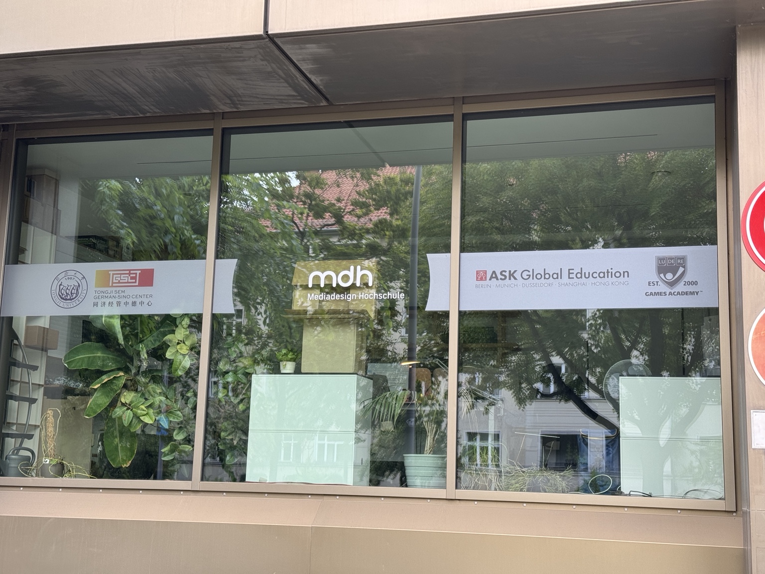 The Mediadesign Hochschule campus window signage in Berlin, with green planting reflected in the glass.