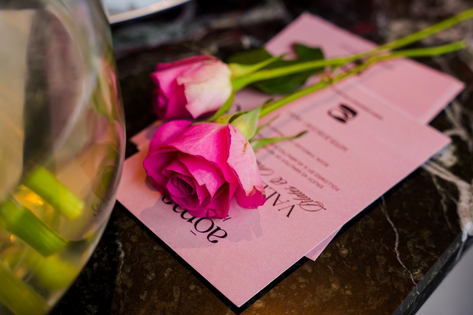 Two pink roses resting on pink aōma Valentine's event cards on a dark marble surface.