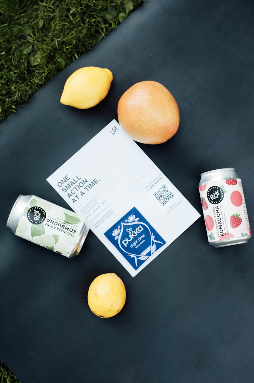 A Living with Mindfulness card reading One small action at a time laid on a yoga mat with kombucha cans, tea and citrus fruit.