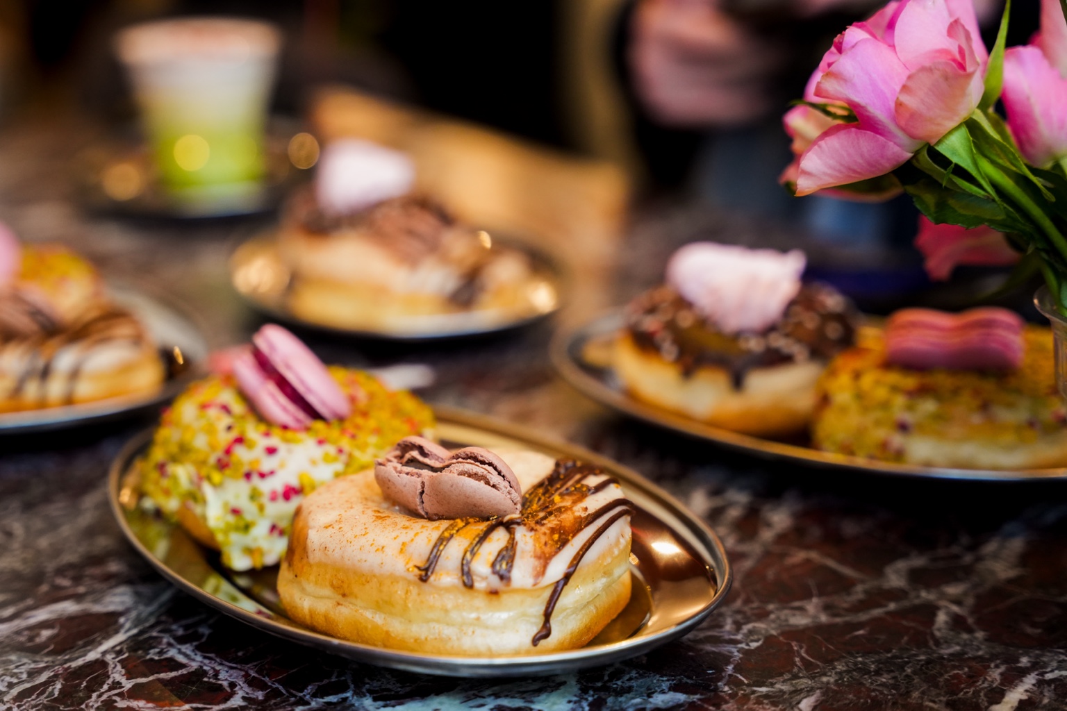 Close-up of Brammibal's Valentine's doughnuts topped with macarons and chocolate drizzle on small metal plates beside pink roses.