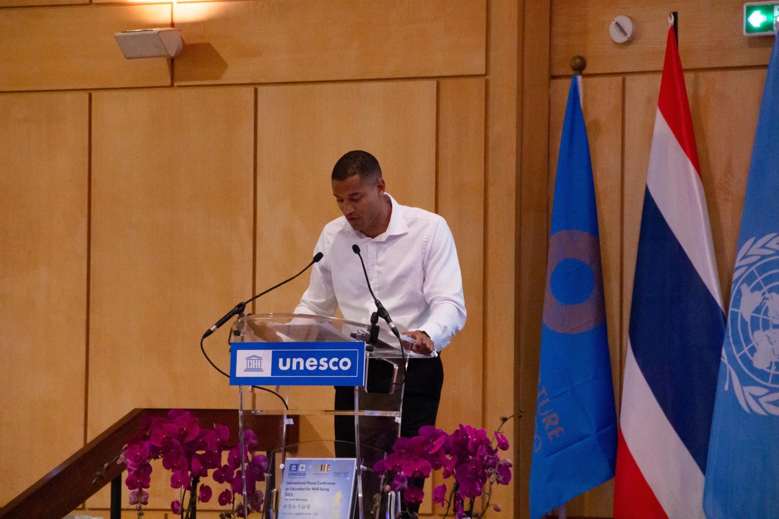 Nathan-Josias Boum speaking from a UNESCO lectern at the International Peace Conference on Education for Well-being, national and UN flags in the background.