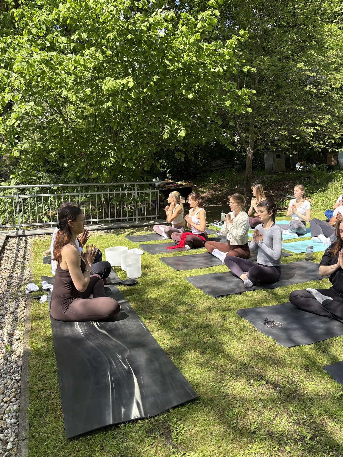 Participants seated cross-legged with hands at heart centre during a garden meditation, crystal singing bowls on a mat nearby.