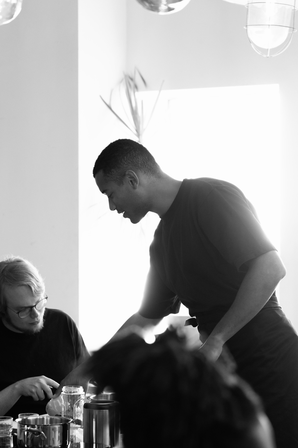 Black-and-white photograph of Nathan leading a mindful cooking workshop, guiding two participants preparing plant-forward ingredients at a shared table.